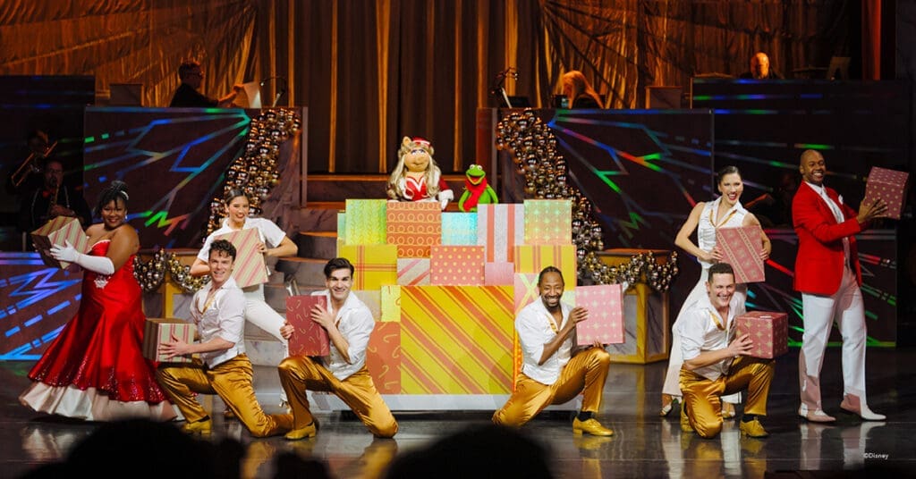 A vibrant holiday stage performance featuring Miss Piggy and Kermit the Frog surrounded by dancers in gold pants, white tops, and red festive attire. The cast holds colorful wrapped presents in front of a backdrop of shimmering gold drapery and dynamic lighting, creating a joyful, high-energy celebration of music and holiday cheer.