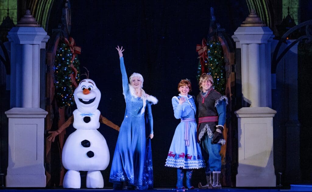 A live stage performance featuring characters from Disney’s Frozen—Olaf, Elsa, Anna, and Kristoff—standing between two decorated pillars with festive garlands and bows. Elsa raises her hand in a joyful pose while the others smile warmly, dressed in their signature winter costumes. The dark backdrop and bright lighting highlight the holiday magic of the scene.