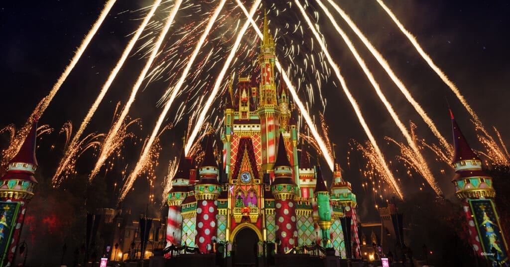Cinderella Castle at Walt Disney World illuminated with vibrant holiday projections in red, green, and gold while fireworks burst dramatically across the night sky. The castle’s towers and spires glow with festive patterns, creating a dazzling celebration of light and color against the dark backdrop.