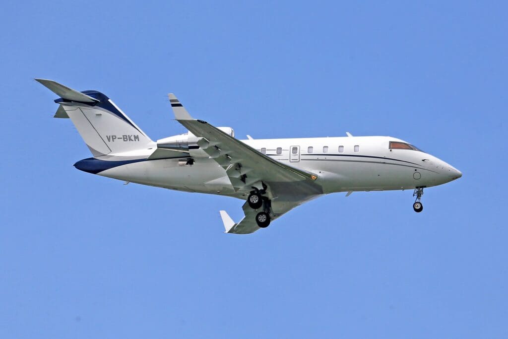 A white private jet with registration number VP‑BKM flying against a clear blue sky, landing gear extended and engines mounted at the rear.