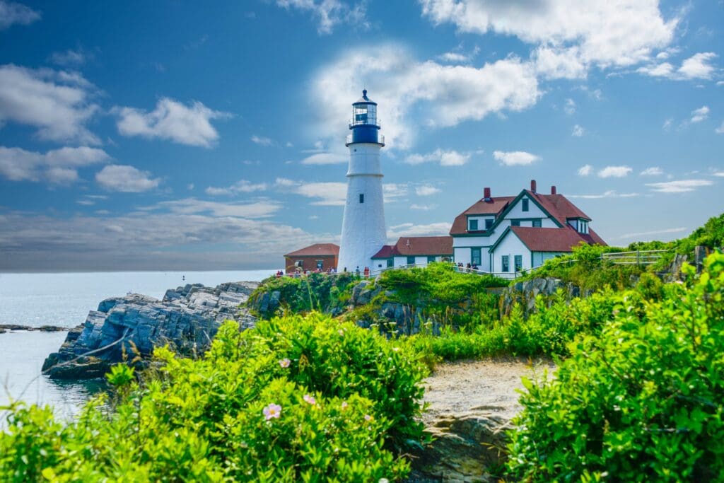 A white lighthouse with a black lantern top stands on a rocky coastline beside red-roofed keeper’s houses, surrounded by lush green vegetation under a bright blue sky with scattered clouds.