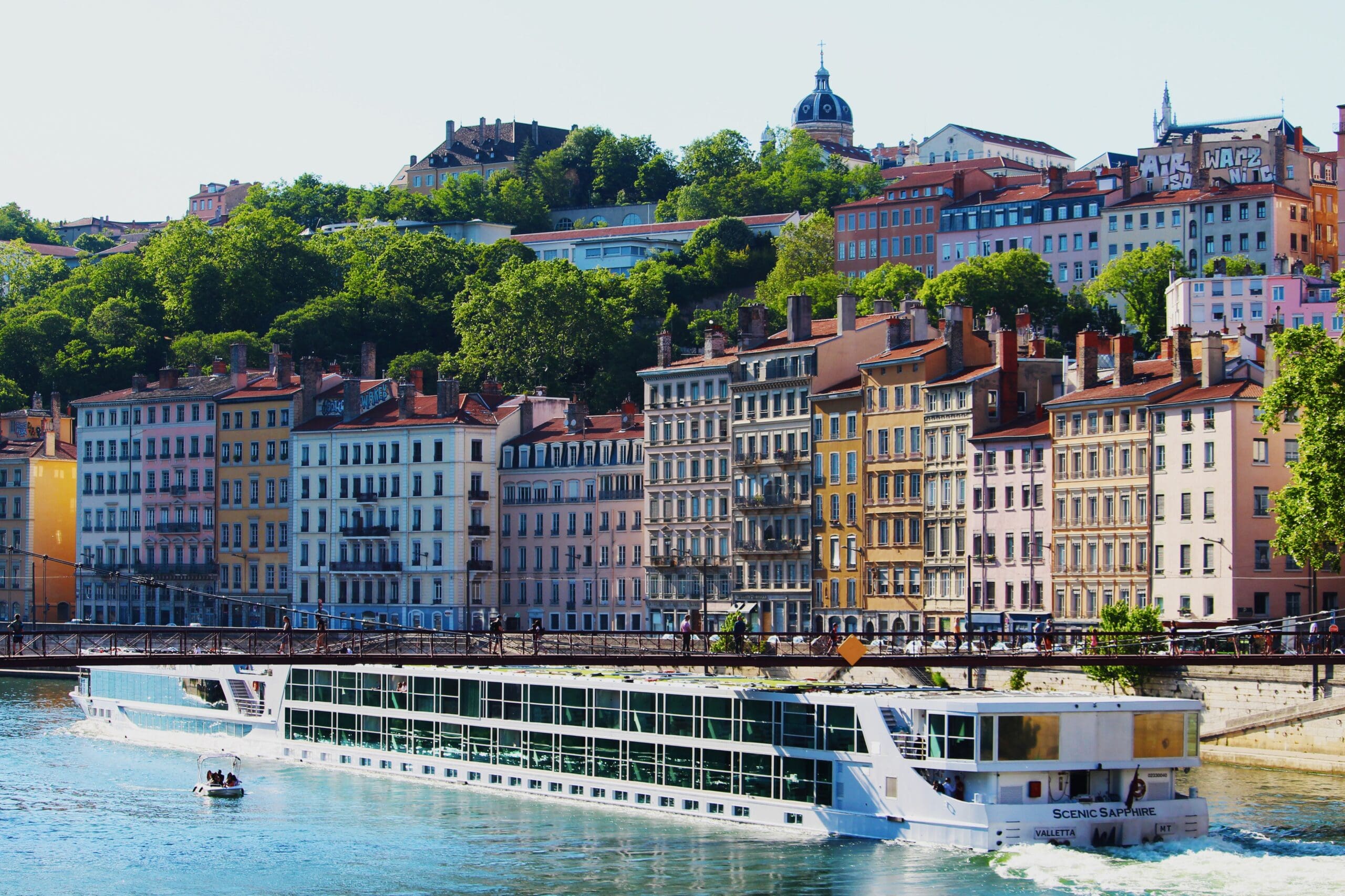 A river cruise ship sailing along the Saône River in Lyon, France, with colorful historic buildings and a tree-lined hillside in the background.