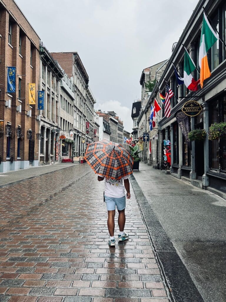 A person holding a plaid orange umbrella walks down a wet cobblestone street lined with historic buildings and colorful international flags on a rainy day.