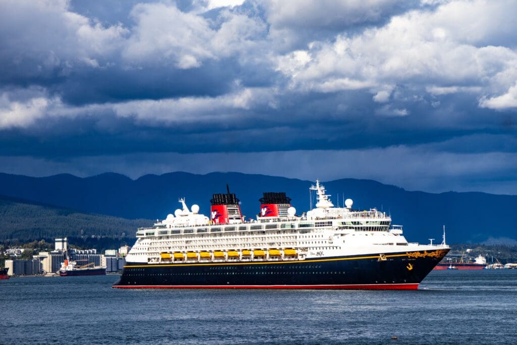 A Disney Cruise Line ship sailing on open water under dramatic storm clouds, with mountains and a coastal city visible in the background.