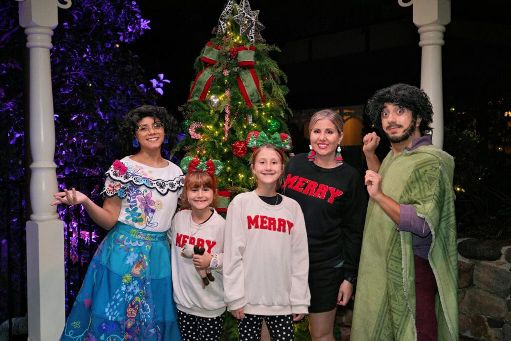 A festive family photo taken outdoors at night featuring five people standing in front of a decorated Christmas tree with red ribbons and a silver star. Two adults are dressed as characters from Disney’s Encanto, while the others wear matching “MERRY” sweatshirts and holiday accessories, creating a joyful blend of Christmas cheer and Disney-inspired fun.