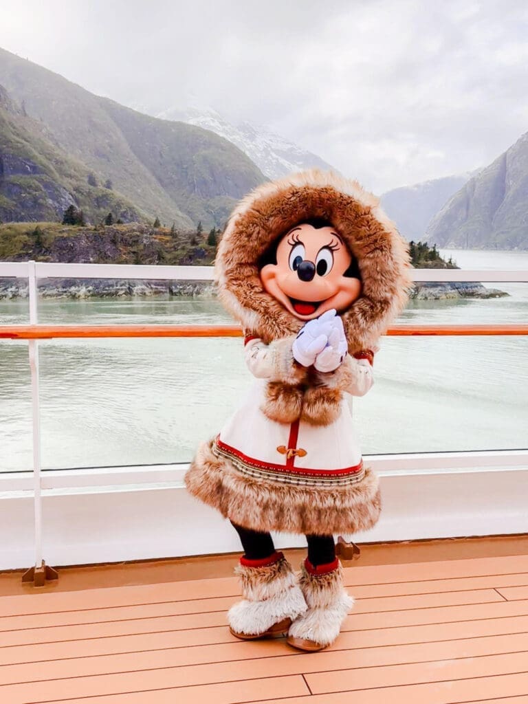 Minnie Mouse dressed in a fur-trimmed winter outfit posing on the deck of a Disney cruise ship sailing through an Alaskan fjord with misty mountains in the background.