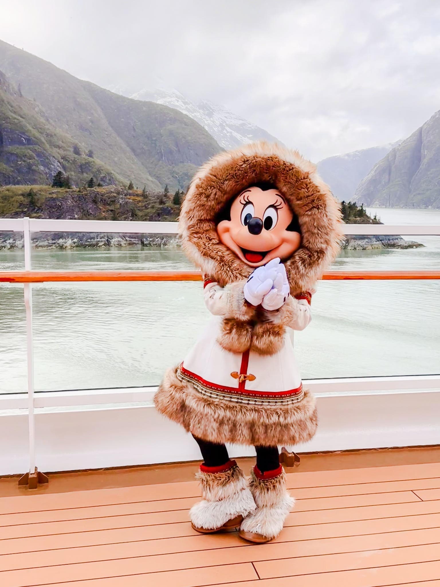 Minnie Mouse dressed in a fur-trimmed winter outfit posing on the deck of a Disney cruise ship sailing through an Alaskan fjord with misty mountains in the background.