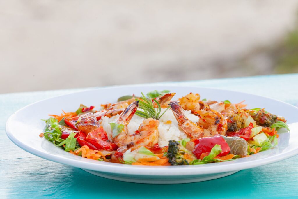 A white plate of grilled shrimp served over a mound of white rice, surrounded by colorful vegetables including red bell peppers, broccoli, shredded carrots, and lettuce, garnished with a fresh herb sprig. The dish sits on a turquoise table with a blurred beach background.