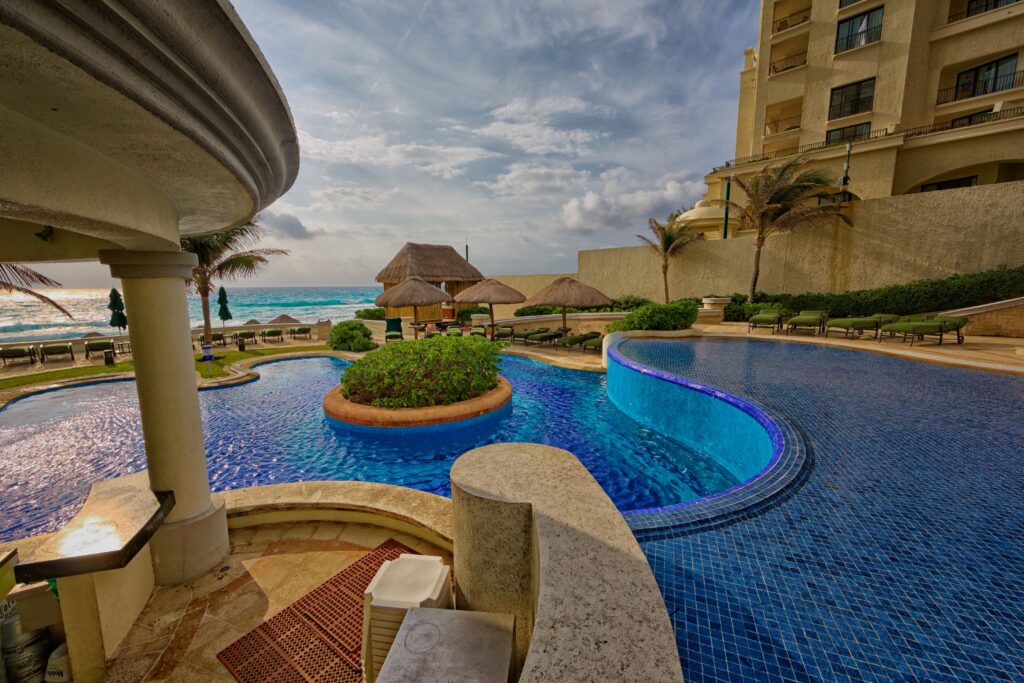 A luxurious oceanfront resort pool with deep blue mosaic tiles and curved edges, featuring a small raised section with a waterfall effect and a circular planter of greenery in the center. Thatched umbrellas and lounge chairs line the poolside, with palm trees and the ocean visible in the background under a partly cloudy sky.
