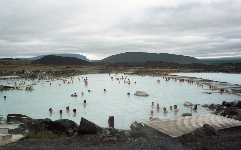 People relaxing in a large geothermal lagoon surrounded by volcanic rocks and distant hills under a cloudy sky.